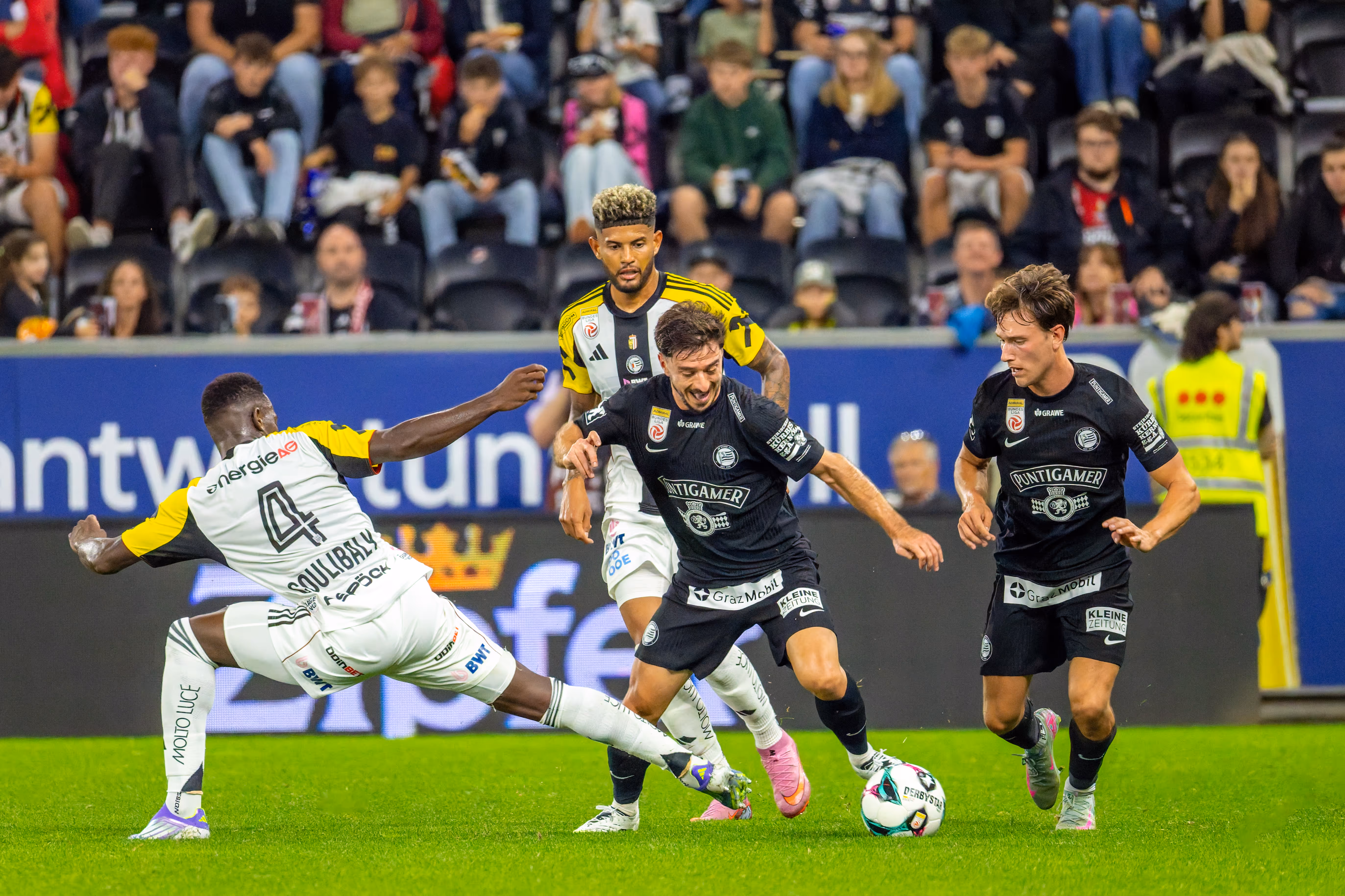 Vier Fußballspieler sind in einen intensiven Zweikampf um den Ball verwickelt, wobei ein Spieler von Sturm Graz den Ball am Fuß führt und ein Spieler des LASK eine Grätsche ausführt