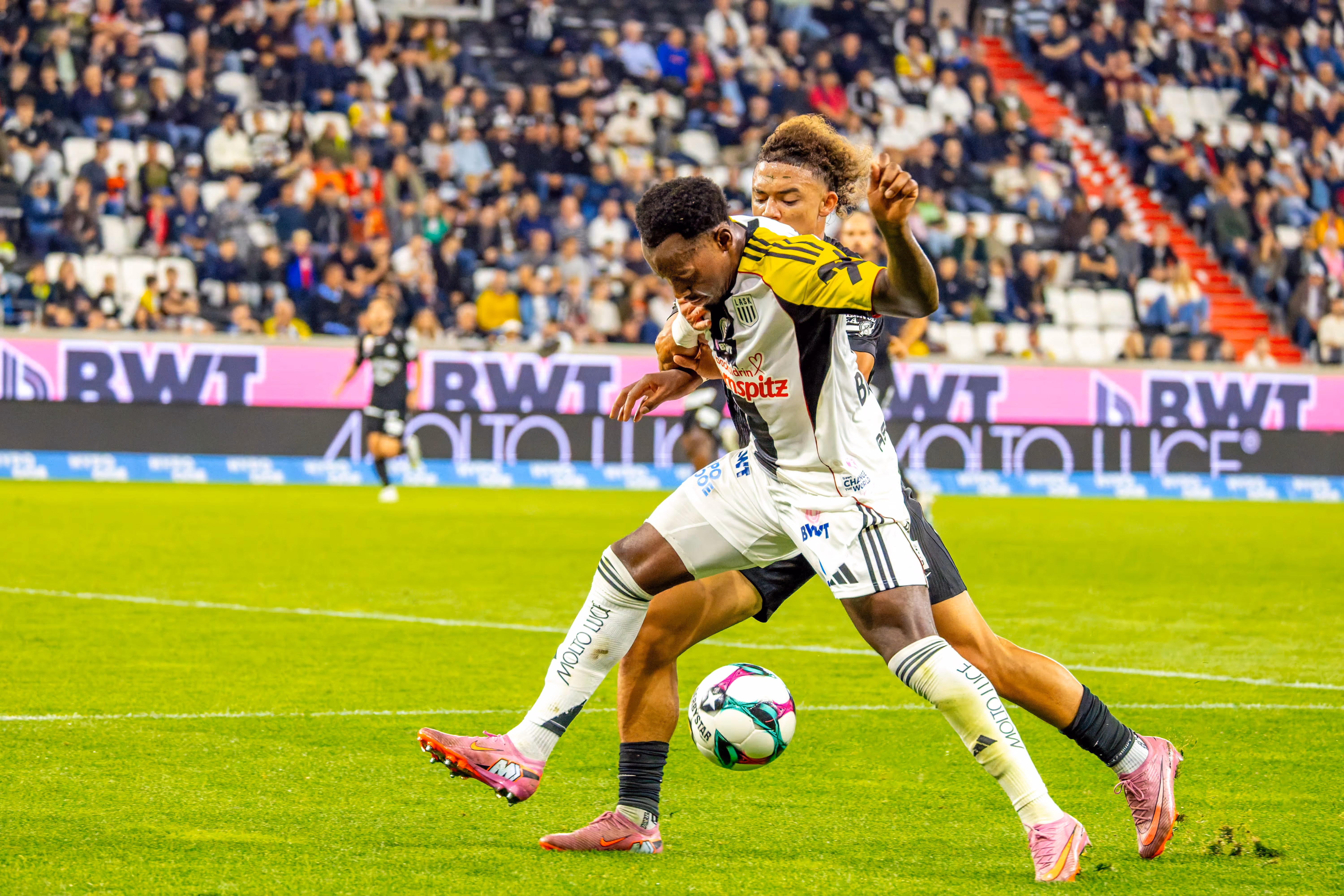 Zwei Fußballspieler in unterschiedlich farbigen Trikots ringen eng um den Ball auf dem Spielfeld vor einer gefüllten Zuschauertribüne