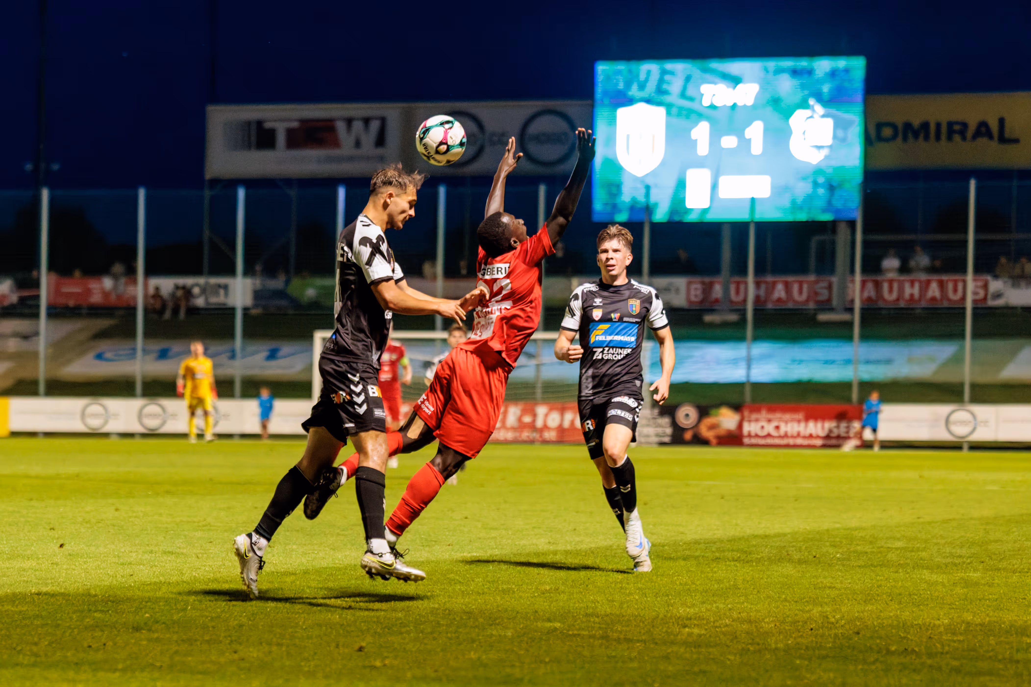 Spieler der SPG HOGO Wels und des SKN St. Pölten liefern sich einen dynamischen Luftzweikampf um den Ball auf dem Fußballfeld unter Flutlicht, während die Anzeigetafel ein 1:1 anzeigt