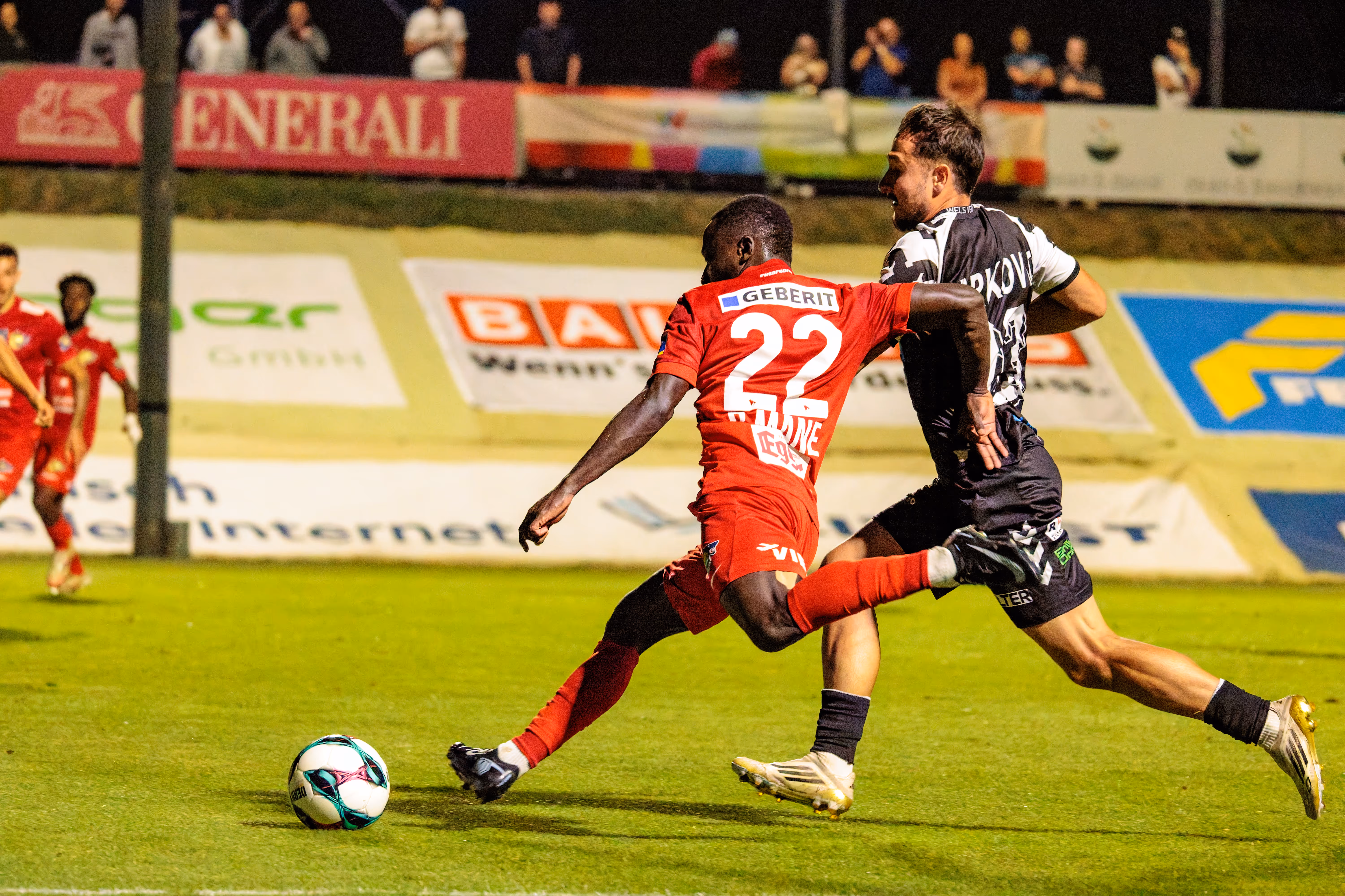 Ein dynamischer Zweikampf am Spielfeldrand zwischen einem Spieler von Herta Wels im roten Trikot mit der Nummer 22 und einem Spieler des SKN St. Pölten im schwarz-weißen Trikot