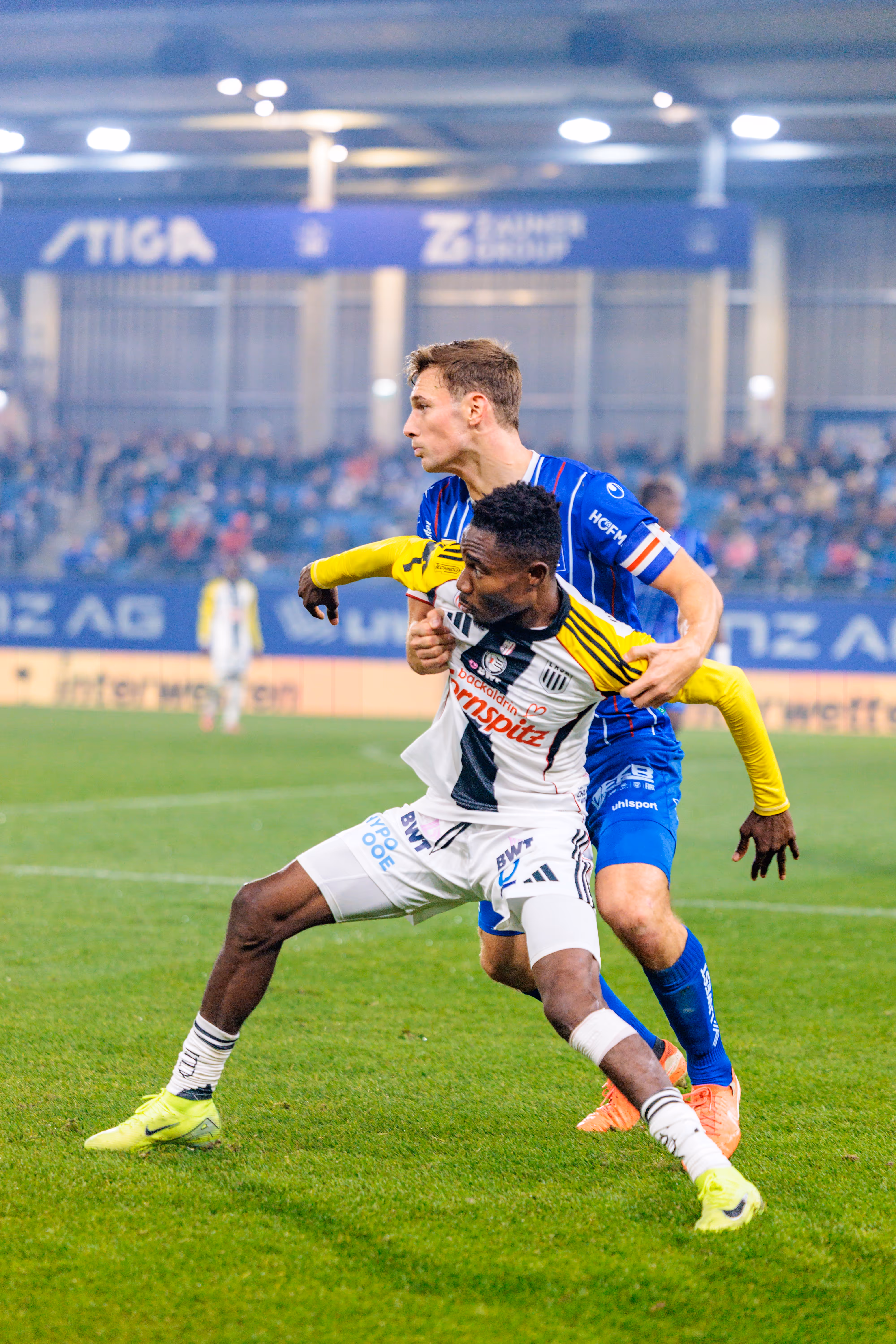 LASK-Spieler George Bello im schwarz-weiß gestreiften Trikot wird von einem Spieler von Blau-Weiß Linz im blauen Trikot in einem intensiven Zweikampf festgehalten