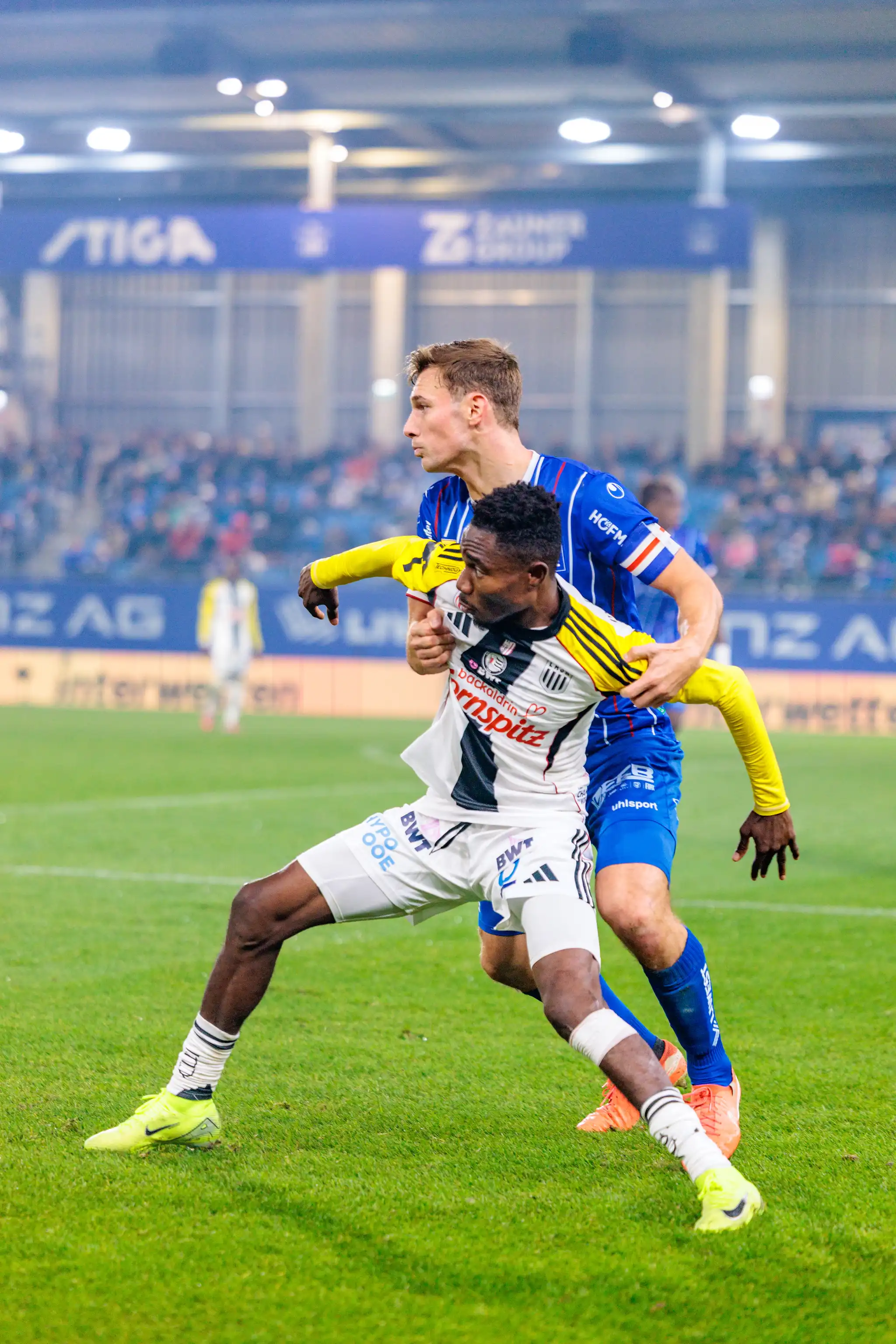 LASK-Spieler George Bello im schwarz-weiß gestreiften Trikot wird von einem Spieler von Blau-Weiß Linz im blauen Trikot in einem intensiven Zweikampf festgehalten