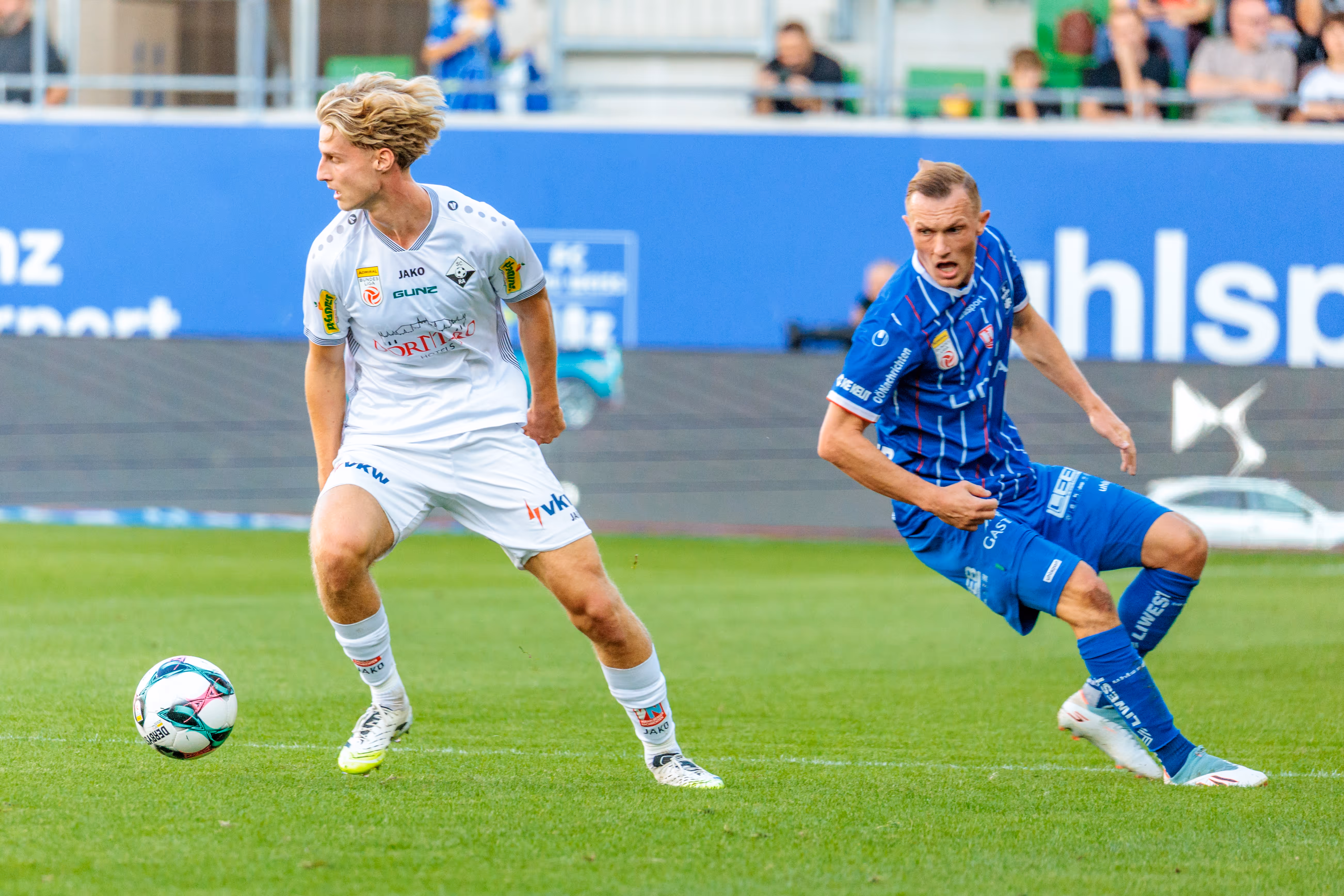 Ein Spieler des SCR Altach im weißen Trikot behauptet den Ball in einem dynamischen Zweikampf gegen einen heraneilenden Spieler von Blau-Weiß Linz