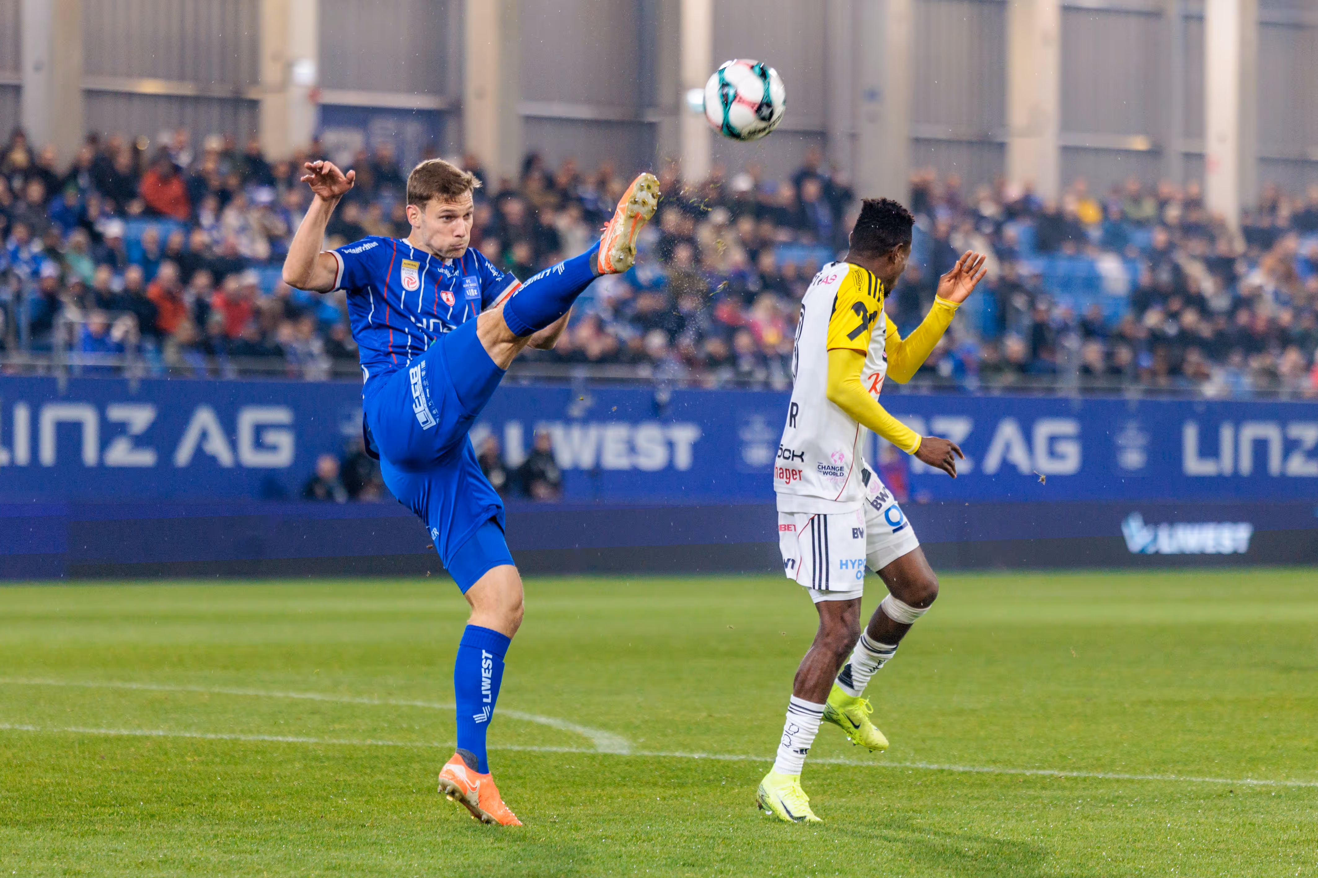 Ein Fußballspieler von Blau-Weiß Linz in blauem Trikot klärt den Ball dynamisch mit einem hohen Tritt, während ein LASK-Spieler in weiß-gelbem Dress danebensteht