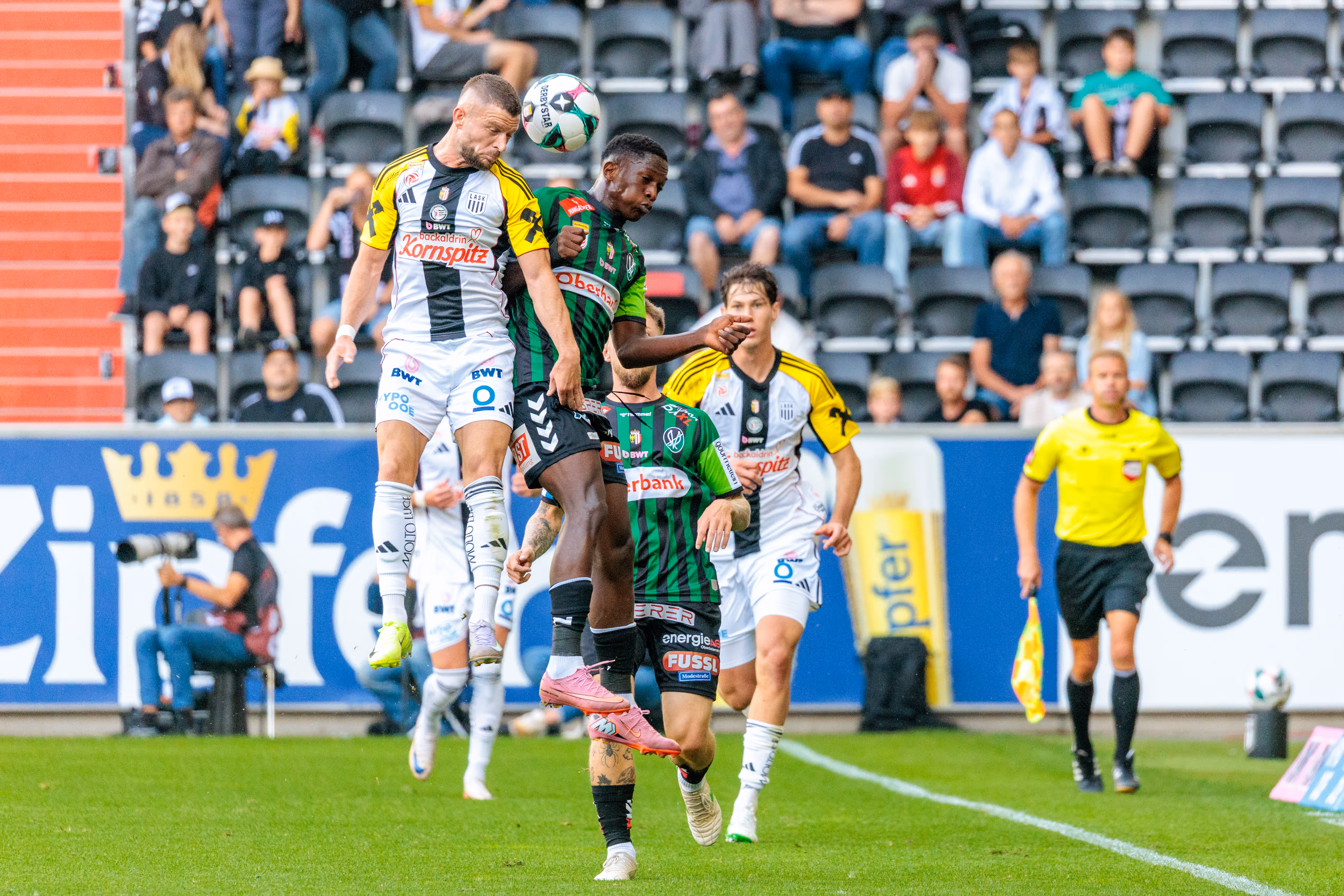 Ein Fußballspieler in einem schwarz-weiß-gelben LASK-Trikot und ein Spieler in einem grün-schwarz gestreiften Trikot der SV Ried springen gleichzeitig für einen Kopfball