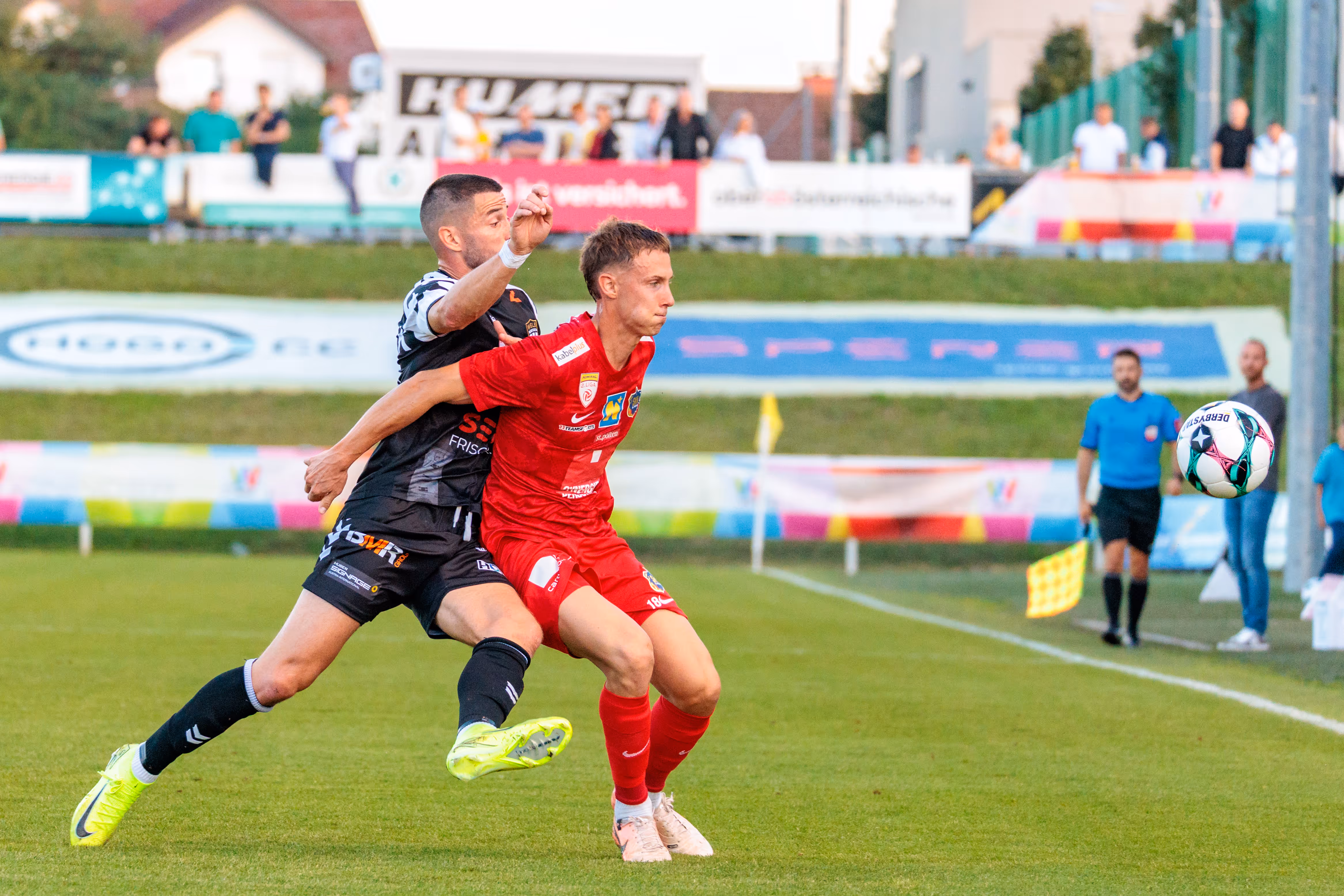 Ein Spieler in rotem Trikot und ein Spieler in schwarz-weißem Trikot ringen auf dem Spielfeld energisch um den Fußball