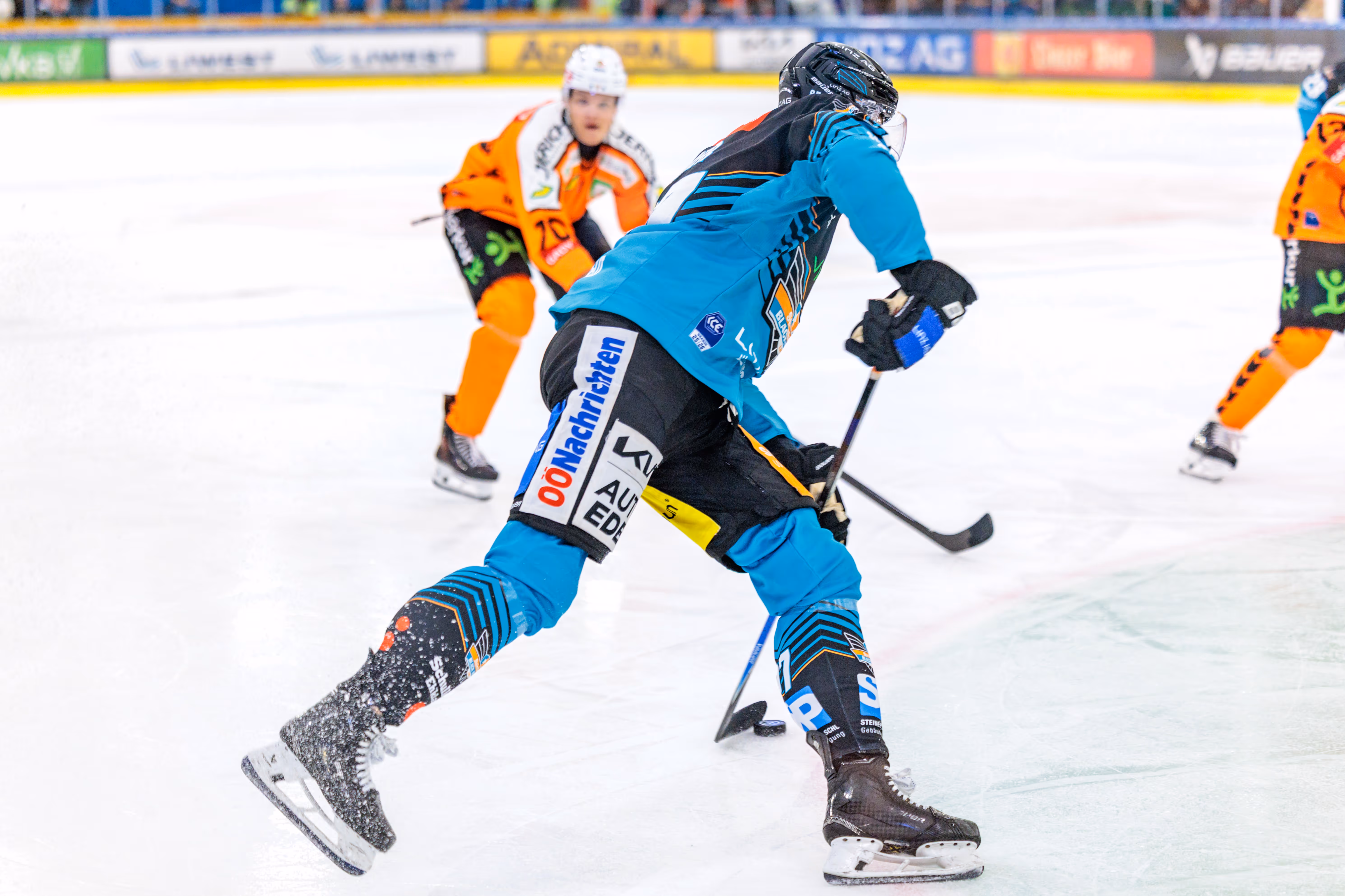 Ein Eishockeyspieler in türkisfarbenem und schwarzem Trikot gleitet über das Eis und erzeugt Eisstaub, während er in einem Zweikampf mit einem orange gekleideten Gegner ist