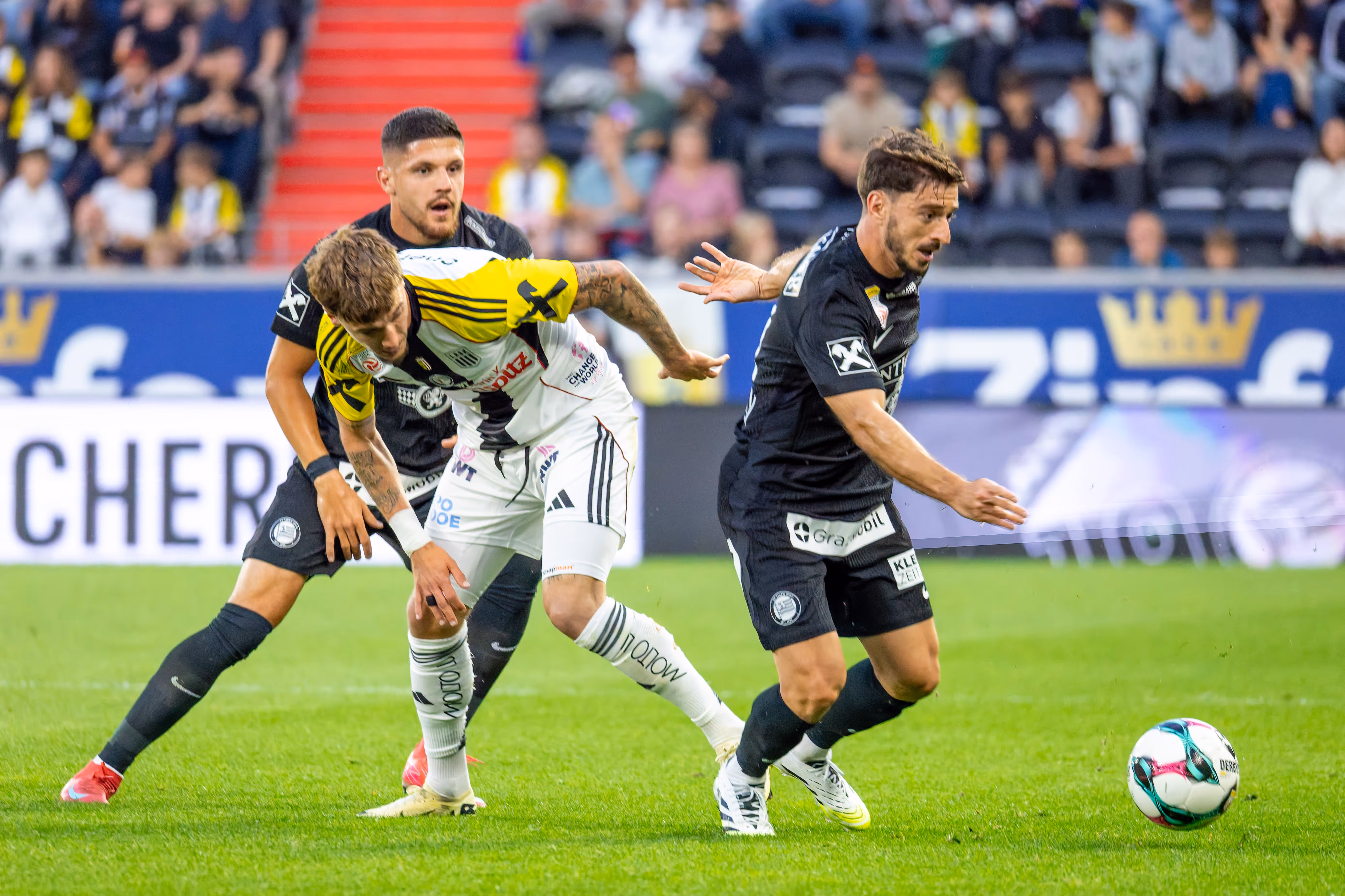 Drei Fußballspieler in unterschiedlichen Trikots kämpfen während eines Spiels im Stadion um den Ball